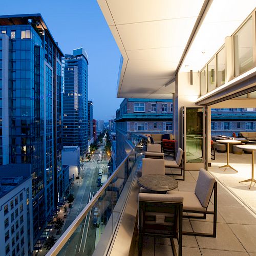 A modern cityscape with a high-rise view, featuring a stylish balcony and furnished seating area overlooking urban buildings and streetlights.