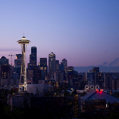 The image shows a skyline view of Seattle at dusk, with the Space Needle and Mount Rainier visible in the background.