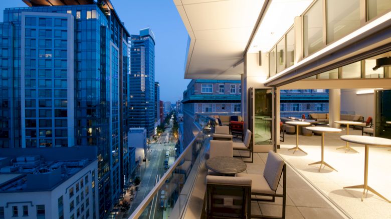 A modern rooftop terrace overlooks a city skyline at dusk with outdoor seating and tables, surrounded by tall buildings.