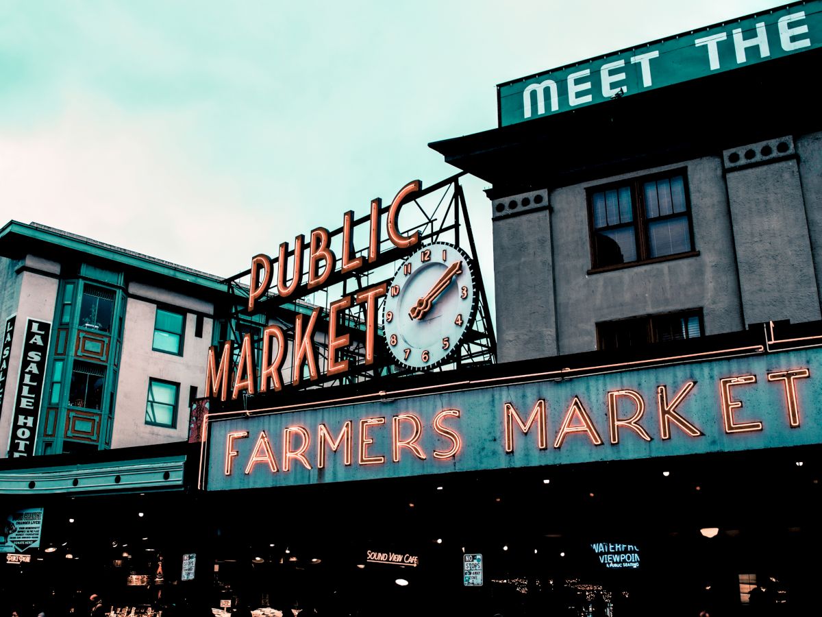 The image shows the exterior of a building with neon signs reading "Public Market" and "Farmers Market" beneath a clock.