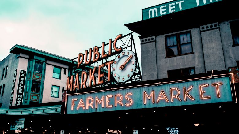 The image shows the exterior of a building with neon signs reading "Public Market" and "Farmers Market" beneath a clock.