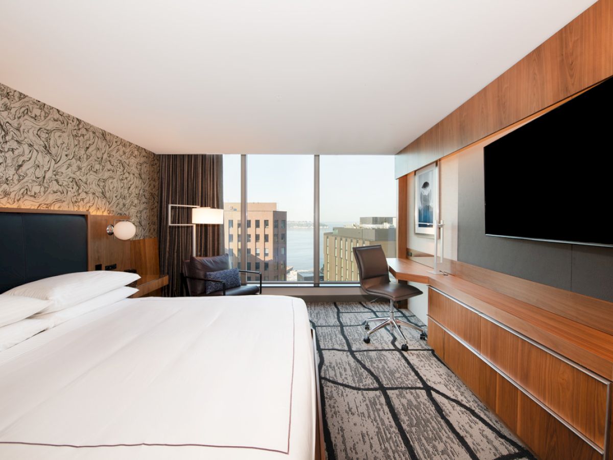 A modern hotel room features a large bed, wall-mounted TV, desk, and city view through large windows.