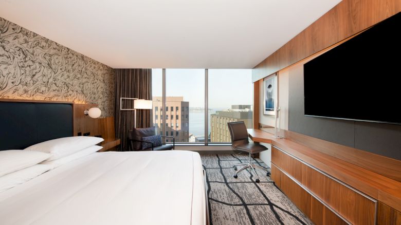 A modern hotel room features a large bed, wall-mounted TV, desk, and city view through large windows.