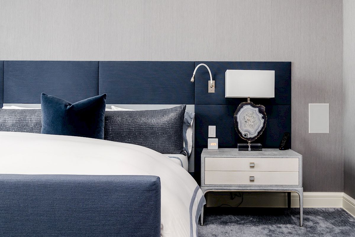 A modern bedroom features a blue upholstered bed, white bedding, a nightstand with a lamp, and a decorative stone.