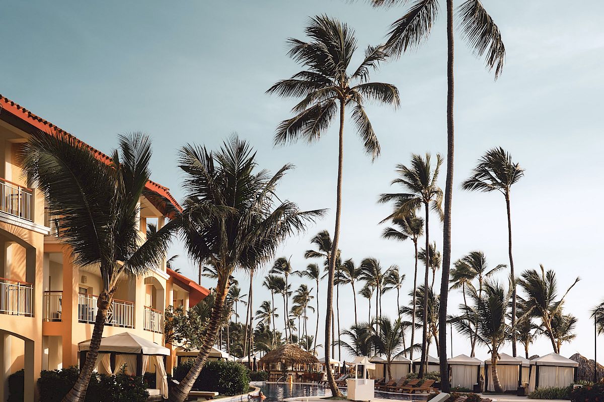 A resort poolside scene with lounge chairs, palm trees, and a building under a clear sky, creating a relaxing tropical atmosphere.