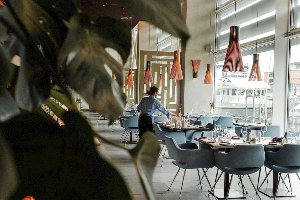 A modern restaurant with elegant decor, featuring blue chairs and large windows; a person is setting tables under stylish orange pendant lights.