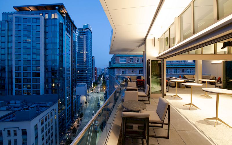 A modern balcony or terrace overlooking a cityscape at dusk, featuring seating areas and tall buildings illuminated by city lights.