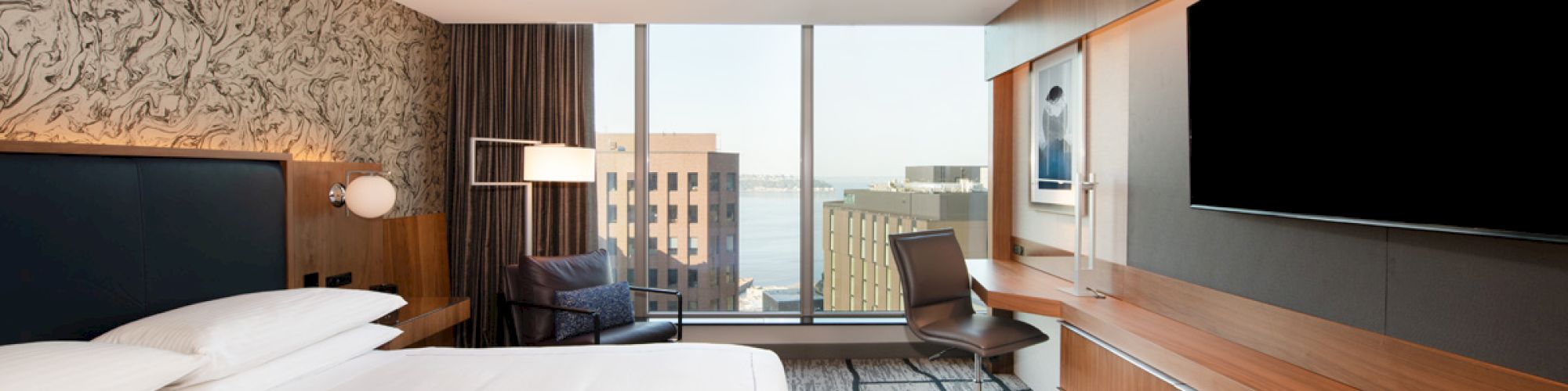 A modern hotel room with a large bed, desk, chair, and big window view. Neutral tones and a large TV add to the contemporary feel.